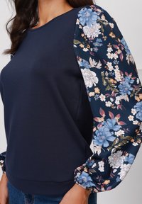 Mørkeblå topp med lange ermer i blomstermønster som har blå og hvite blomster, elastiske mansjetter og en glatt, tettsittende kropp.