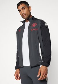 Chaqueta de chándal negra con el logo rojo del Manchester United, detalles en blanco, cremallera en la parte delantera y puños elásticos. Fabricada con material ligero y transpirable.