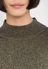 Halsringad, ribbad stickad topp i mörkgrön med glittrande guldtrådar. Närbild som lyfter fram textur och kragdesign.