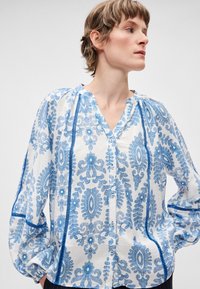 Λευκό πουκάμισο με μπλε λουλουδάτο σχέδιο, κουμπιά μπροστά, V-λαιμόκοψη και φουσκωτά μανίκια. Διαθέτει μπλε λεπτομέρειες στο τελείωμα κατά μήκος των ραφών και των κουμπιών.