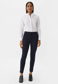 Witte knoop blouse en donkerblauwe slim-fit broeken met een gladde textuur, gecombineerd met glanzende zwarte loafers.