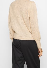 Beige neulepaita, jossa on rento istuvuus ja teksturoitu kangas; siinä on pitkät, hieman puffimaiset hihat ja resorireuna vyötäröllä.