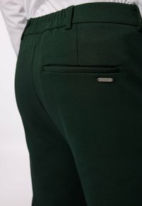 Gröna byxor i glatt tyg, med elastisk midja, en bakficka och en liten metallmärkning med "JP 1880" på.