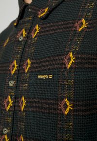 Wrangler-ruutupaita tummanvihreänä, ruskeilla ja keltaisilla yksityiskohdilla. Varustettu nappikiinnityksellä ja kudotulla, hienovaraisella tekstuurilla.