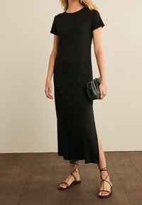 Robe noire midi à manches courtes, avec une silhouette droite et une fente sur le côté. Associée à des sandales noires à brides et portant un petit sac noir.