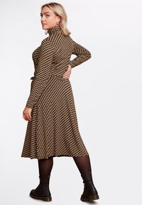 Vestito marrone a fantasia con maniche lunghe e colletto alto. Lunghezza sotto il ginocchio con vita aderente e gonna a campana. Stivaletti neri alla caviglia.