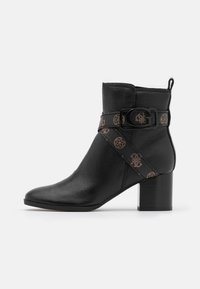 Bottine en cuir noir avec un bout pointu, un talon carré et une sangle décorative arborant un motif de logo. Texture lisse avec des détails de couture.