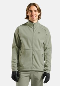 Grüne Softshell-Jacke mit hohem Kragen, Reißverschluss vorne, zwei Seitentaschen und schwarzen Handschuhen; glatte Textur und minimalistische Markenakzente.