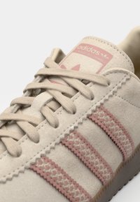 Krem-bijele Adidas tenisice s ružičastim prugama, od suède materijala, teksturirane površine i tradicionalnog dizajna s vezicama. Sadrži logotip na jeziku.
