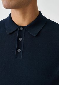 Mereväe sinine polo särk soonikust kangast, millel on klassikaline krae ja kolme nupuga plastron sobivate nuppudega.