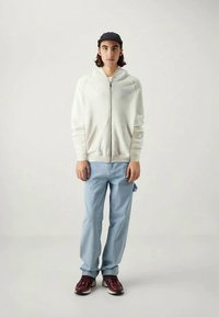 Gaiši pelēks izsistā jaka ar ribotām aprocēm, uzvilkta virs zilām džinsiem. Melna cepure un bordo kedas papildina apģērbu. Minimālisma dizains.