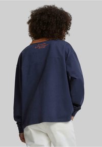 Marineblå langermet sweatshirt med brun ribbestrikket krage og oransje brodert tekst på ryggen, laget av myk bomullsstoff.
