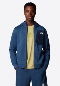 Marineblauwe zip-up hoodie met een voorzak, gecombineerd met een lichtgele t-shirt met een berglogo, gedragen met bijpassende marineblauwe shorts.