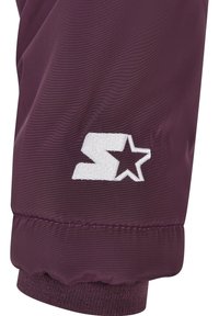 Gros plan sur la manche d'une veste bordeaux avec un poignet élastique et un logo brodé blanc "S" et étoile près du poignet.