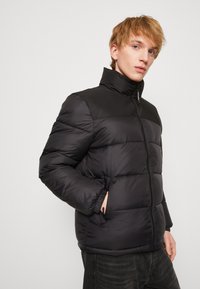 Schwarze gepolsterte Jacke mit hohem Kragen und Reißverschluss. Verfügt über ein gestepptes Muster, elastische Bündchen und seitliche Taschen für Funktionalität.