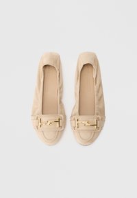 Beežidest seemisnahast kahe kuldse metallist pandlaga mokassiini, millel on ruudukujuline ninaosa ja pehmendatud sisetallad, helehalli tausta peal.