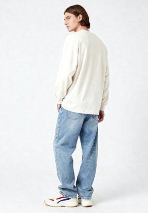 Junge Person mit braunen Haaren, die ein langärmeliges weißes Hemd, lockere hellblaue Jeans und weiße Sneakers mit roten und blauen Details trägt.