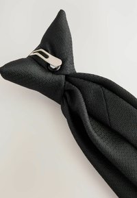 Bandeau en tissu noir avec une pointe triangulaire, surface texturée et un détail en métal argenté pour le fermoir.