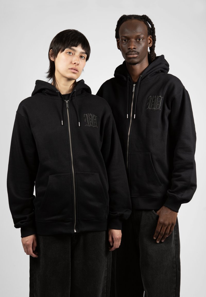 Sudaderas con cremallera en color negro, de mezcla de algodón, que cuentan con bolsillos frontales y acentos de logotipo bordado. Ambos modelos llevan pantalones oscuros y sueltos.