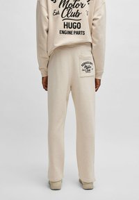 Beige sweatpants hebben een achterzak met de tekst "SERVICE DEPT. Motor Club". De stof is zacht en textuurrijk, wat zorgt voor een relaxte pasvorm.