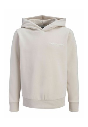 Dreng med krøllet hår, smilende, iført en beige hættetrøje, blå jeans og hvide sneakers, stående mod en ensfarvet hvid baggrund.