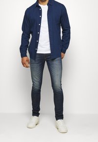 Mörkblå knapp-up skjorta över en vit t-shirt, kombinerad med slim-fit mörkblå jeans och vita sneakers, allt mot en neutral bakgrund.