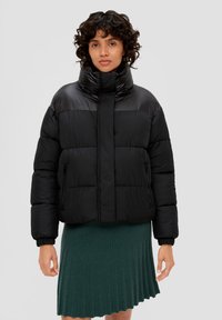 Schwarze Steppjacke mit hohem Kragen, abgestepptem Design, seitlichen Reißverschlusstaschen und elastischen Bündchen, kombiniert mit einem grünen Ripprock.