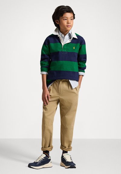 Ριγέ ράγκμπι μπλούζα σε ναυτικό μπλε και πράσινο με λευκό γιακά, χακί παντελόνι και ναυτικές αθλητικές παπούτσια με ανοιχτόχρωμες λεπτομέρειες. Μοντέλο που στέκεται σε ουδέτερο φόντο.