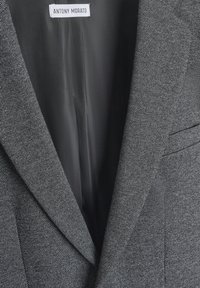 Blazer gris ajusté avec un tissu texturé, des revers nets et une doublure intérieure lisse. L'étiquette indique "ANTONY MORATO."