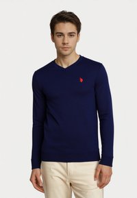 Nuori mies, jolla on päällään tummansininen V-kaula-aukkosweatshirt, jossa on punainen logo rinnassa, ja vaaleat housut, seisoo yksinkertaista valkoista taustaa vasten.