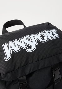 Svart ryggsekk med stripete tekstur, med en stor hvit "JANSPORT"-logo på klaffen og spennehardware for lukking.