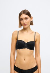 Čierna čipkovaná podprsenka s tvarovanými košíčkami, jemným kvetinovým vzorom, vlnitými okrajmi a nastaviteľnými ramienkami. Mäkká textúra a kostícová podpora.