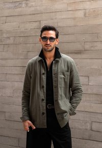 Olivgrüne knöpfbare Jacke mit einem schwarzen Kragen, strukturierter Stoff, einzelne Brusttasche, getragen über einem dunklen Reißverschlusshemd und schwarzen Hosen.