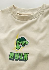 Beige T-shirt met groene geborduurde Hulk-afbeelding, een geel bliksemeffect en gedurfde, gestructureerde "HULK"-tekst in groen eronder.