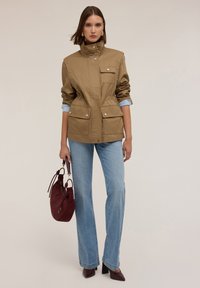 Giacca khaki con bottoni a pressione, due tasche frontali e colletto alto, abbinata a jeans azzurri e una borsa bordeaux.