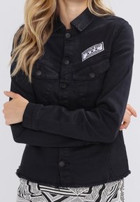 Giubbotto di jeans nero con colletto, chiusura a bottoni, tasche sul petto, orlo sfrangiato e una toppa bianca con logo sul lato sinistro superiore.