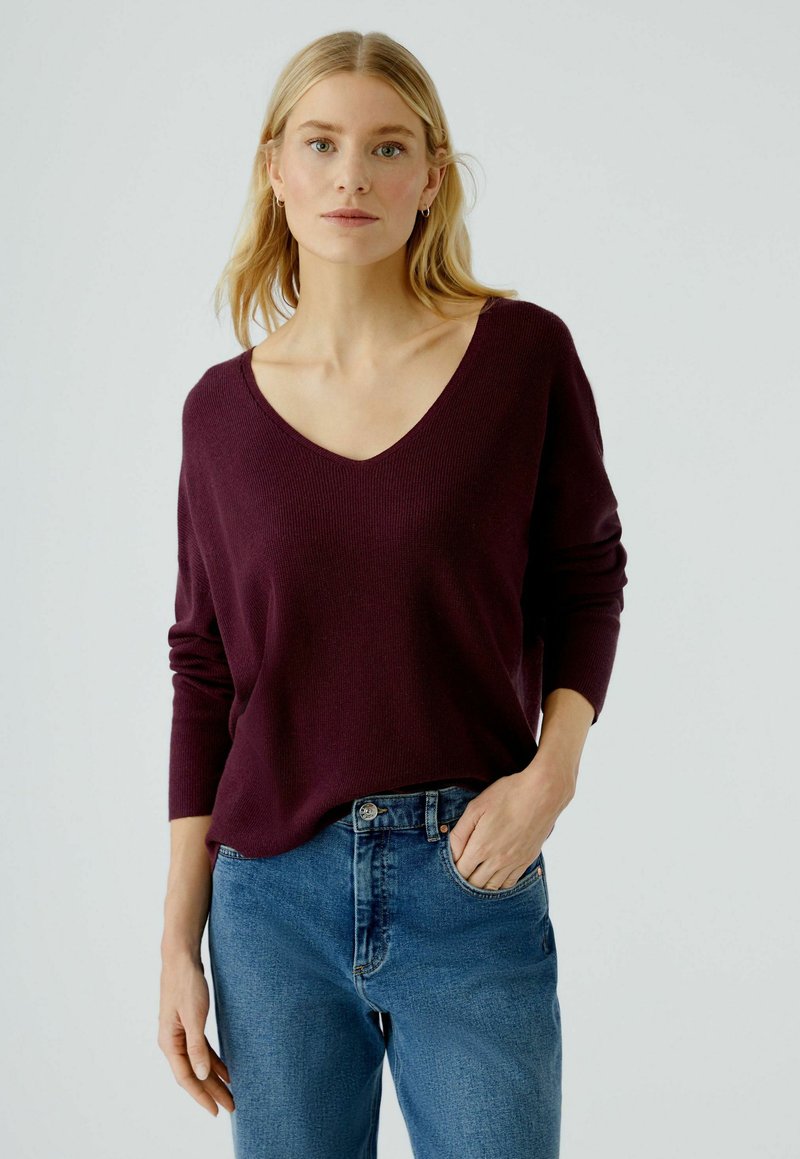 Burgunderfarbener V-Ausschnitt-Pullover mit gerippter Textur und langen Ärmeln, kombiniert mit blauen, hochgeschnittenen Jeans, die einen lässigen Look kreieren.