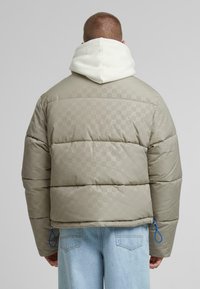 Polstret jakke i lys beige med en subtil mønstret overflade. Har en hvid hætte og blå detaljer ved kanten, med et afslappet fit design.