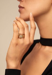 Mano de mujer con los dedos cerca de la cara, llevando anillos de oro apilados con un pequeño charm en forma de corazón en el dedo medio, vistiendo una blusa negra con un collar tipo choker.