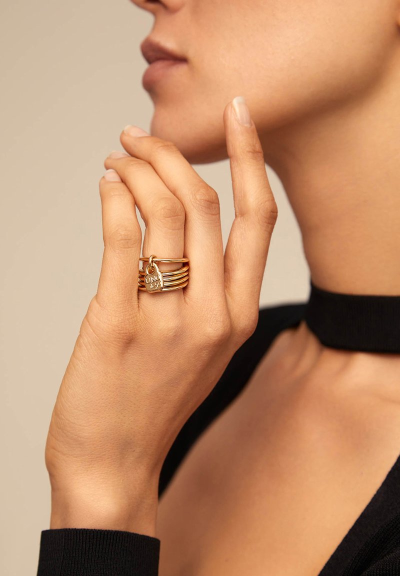 Mano de mujer con los dedos cerca de la cara, llevando anillos de oro apilados con un pequeño charm en forma de corazón en el dedo medio, vistiendo una blusa negra con un collar tipo choker.