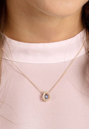 Collier chaîne en or avec un pendentif en forme de larme orné d'une pierre bleue entourée de petites pierres claires, porté sur un haut rose.