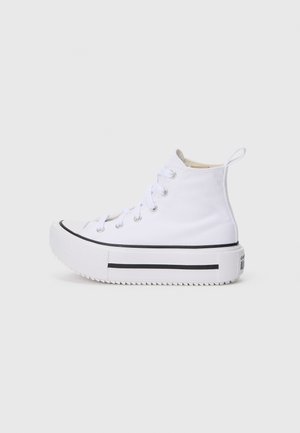Zapatilla blanca de caña alta con suela gruesa de plataforma, franja lateral negra y cordones blancos sobre un fondo liso gris claro.