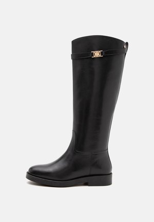 Botte en cuir noire montant jusqu'au genou avec un petit talon, un bout rond et une sangle décorative avec boucle dorée près du bord supérieur.