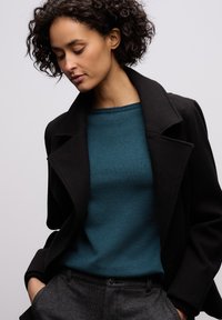 Czarny płaszcz z klapami i wycięciem kołnierza na zielonym swetrze. Do stroju dołączono ciemnoszare spodnie wykonane z tkaniny o teksturze.