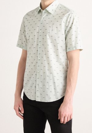 Homme portant une chemise à manches courtes gris clair avec un petit imprimé de requins noirs et un pantalon noir, debout devant un fond uni.