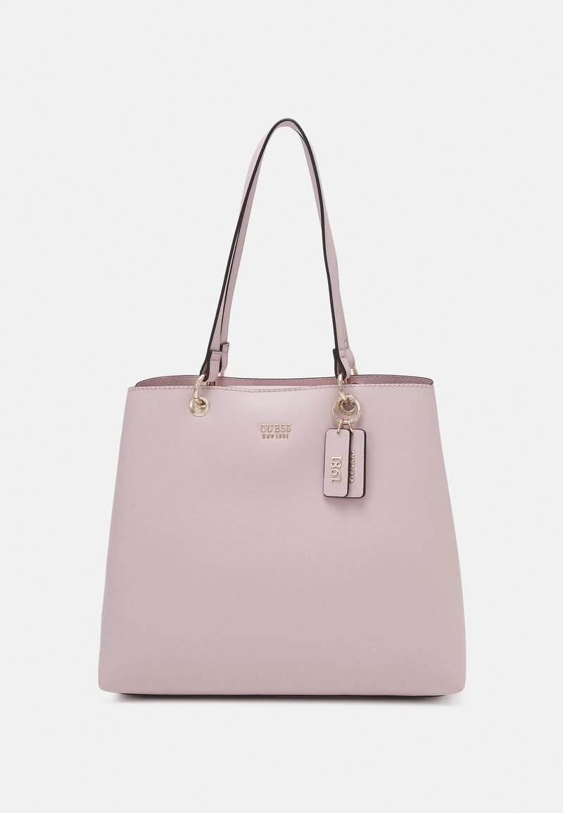 Light pink rectangular handbag with two thin handles, gold accents, and attached tags reading "1981" and "GUESS" on plain background.