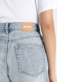 Gros plan d'une personne portant un jean en denim bleu clair avec une étiquette en cuir "WEEKDAY" sur la ceinture et un t-shirt blanc rentré dedans.