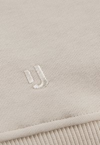 Svetlo béžová tkanina s ripovanou textúrou s vyšívaným logom "J" v zhodnej farbe. Čistý, moderný dizajn s jemným detailom.