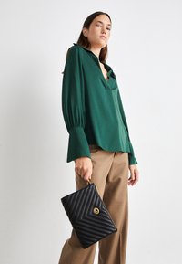 Blusa verde de corte relajado con cuello, combinada con pantalones marrones de talle alto. Clutch negro acolchado con herrajes dorados.