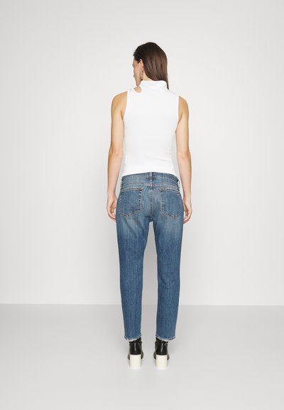 Hvit ermeløs topp med høy hals og utskjæringsdetaljer, paret med blå denim jeans med avslappet passform og frynset kant.